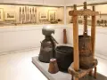 W muzeum Ateny: zabytkowy alembik, kadź i prasa do winogron. W tle ekspozycja narzędzi i obrazów.