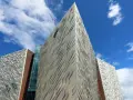 Nowoczesna bryła Muzeum Titanica w Belfaście, z geometrycznymi panelami i szklanymi elementami, pod błękitnym niebem.