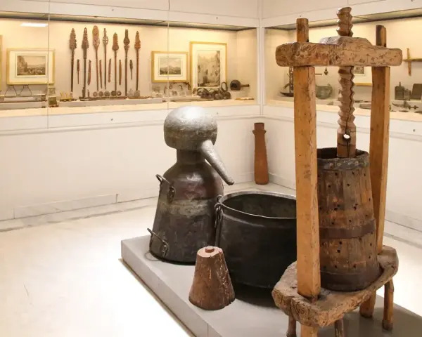 W muzeum Ateny: zabytkowy alembik, kadź i prasa do winogron. W tle ekspozycja narzędzi i obrazów.